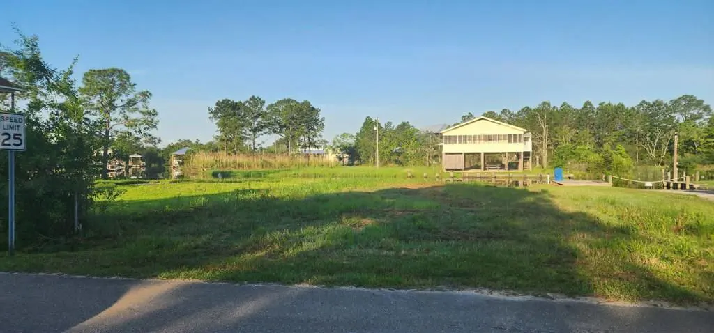 A vacant lot with green grass and trees under a blue sky. A yellow house on stilts is in the background. A speed limit sign is on the left.