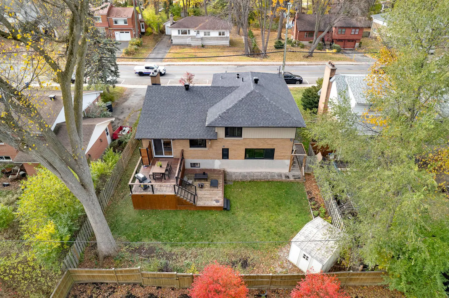 Aerial view of a two-story brick house with a gray roof, a wooden deck with furniture, a green lawn, and a white shed in the backyard.