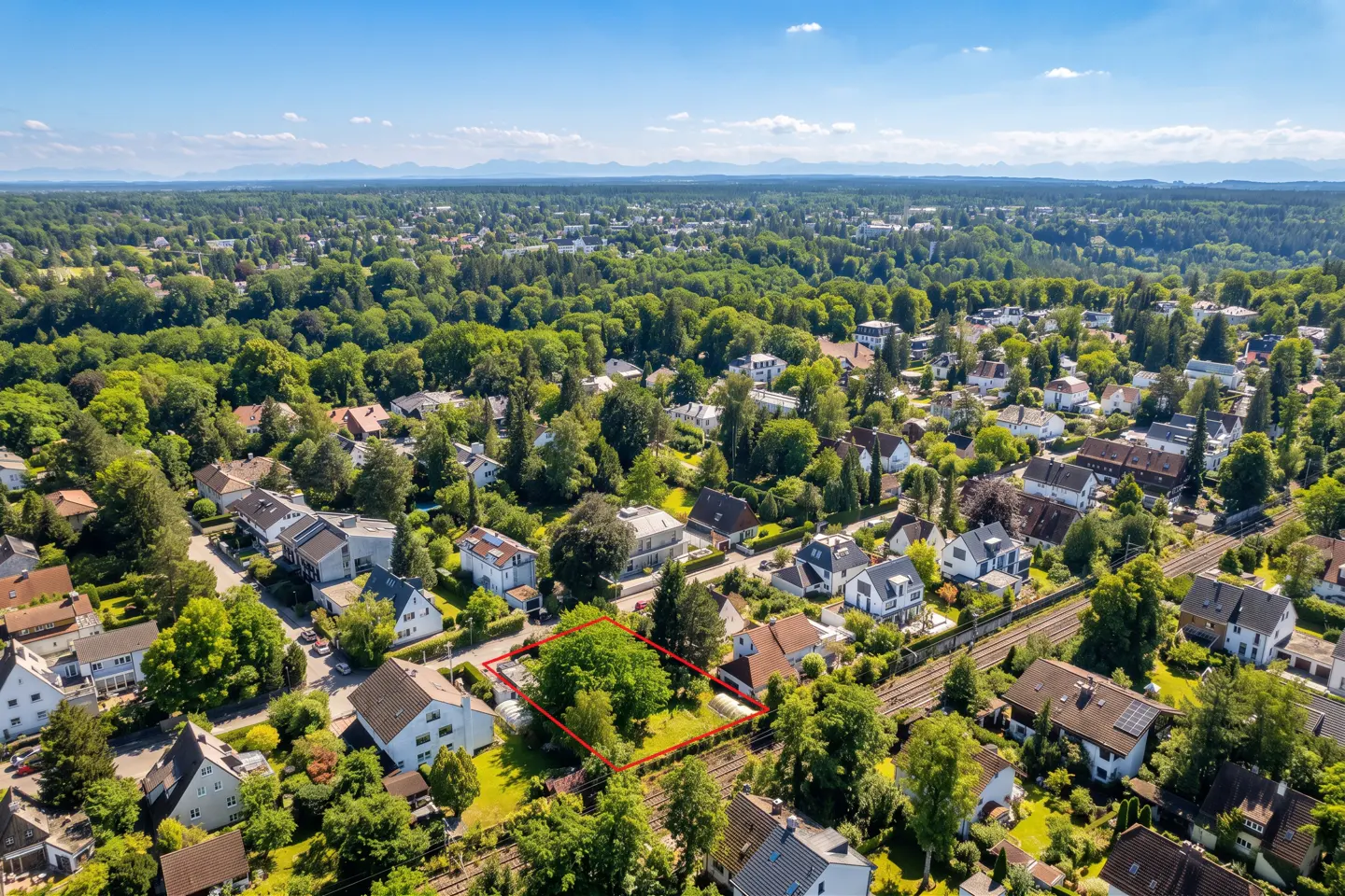 Aerial view of a property outlined in red, surrounded by green trees and houses in a suburban neighborhood. Blue sky and distant mountains.