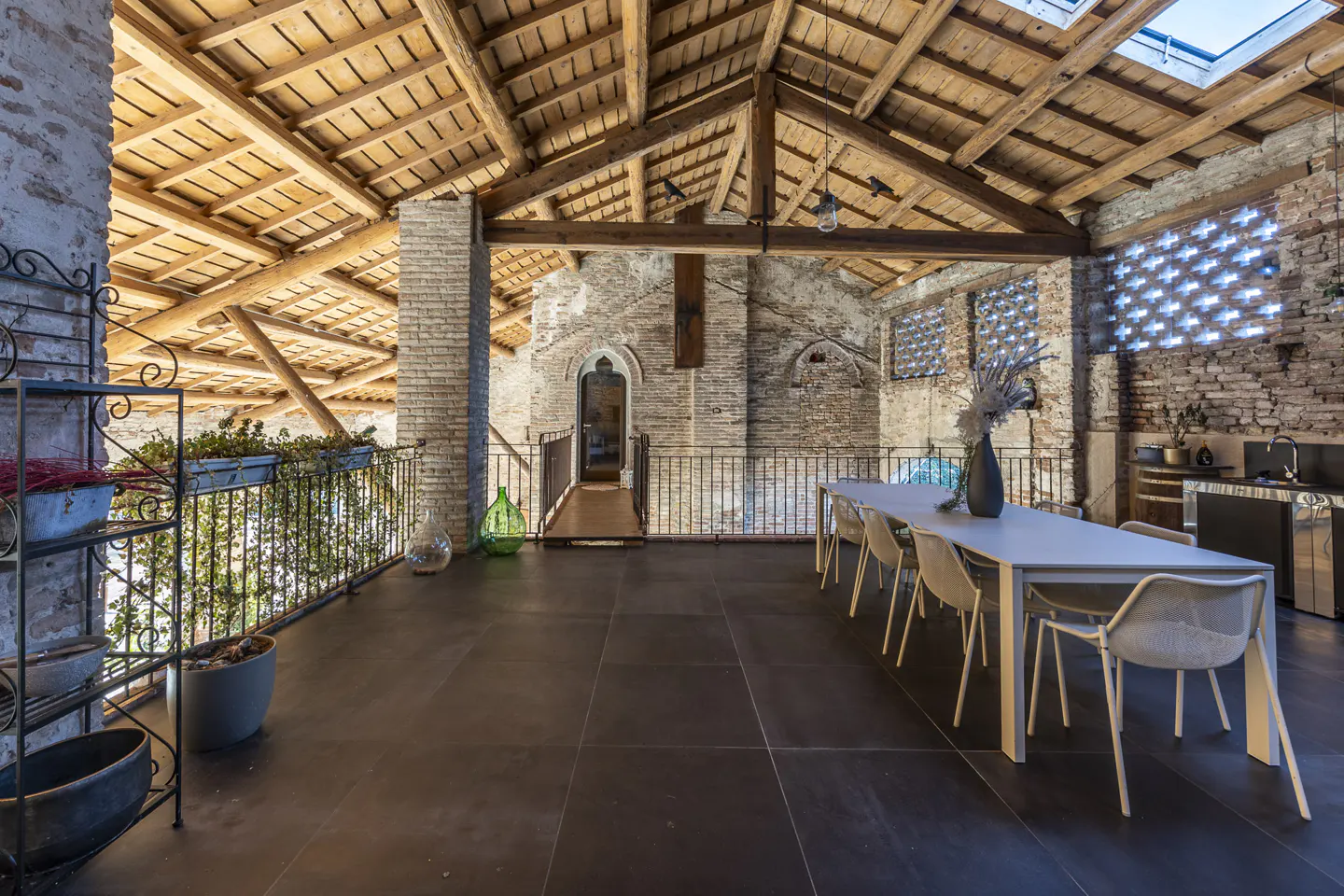 Rustic attic loft with exposed wooden beams, brick walls, dark tile floors, a dining table, and a metal balcony railing.