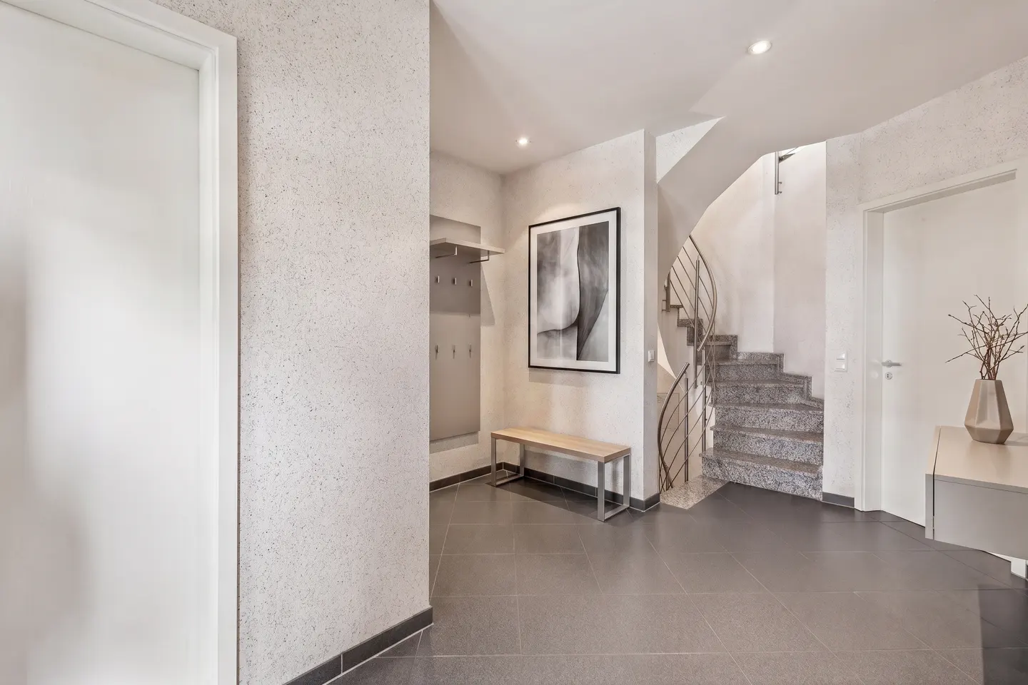 A modern foyer with gray tile floors, white walls, and a spiral staircase. A bench and coat rack are on the left, and a vase with branches is on the right.