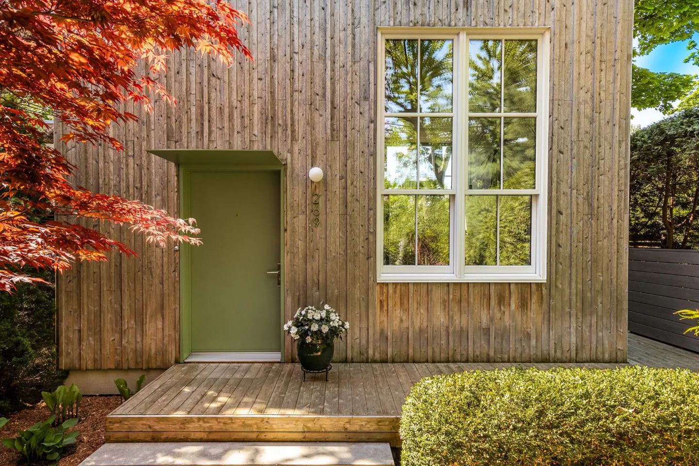 Exterior of a wood-clad house with a green door, white window, and red maple tree.