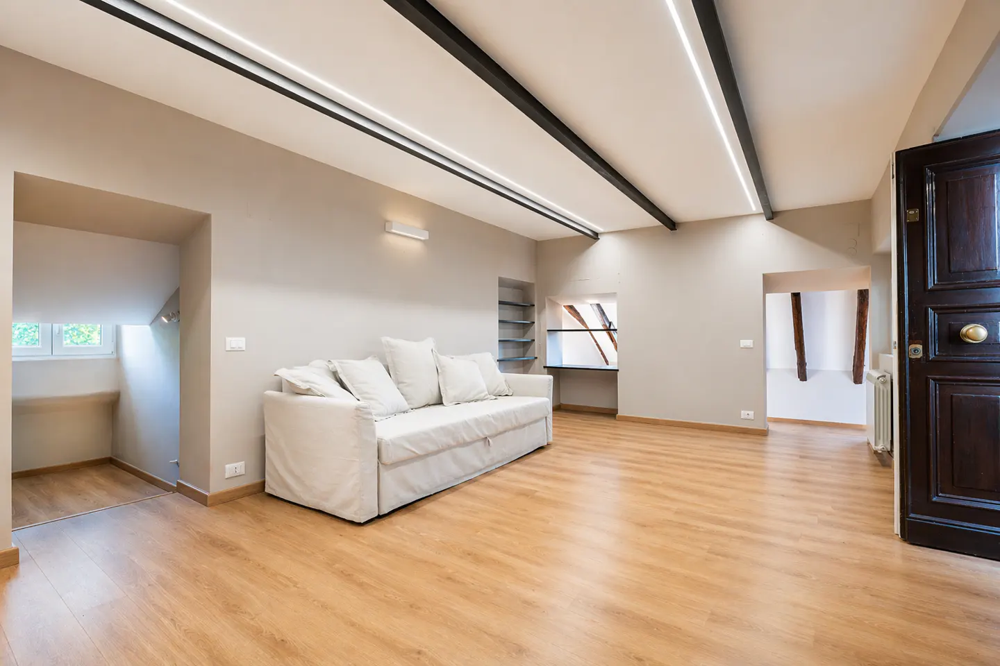 Bright, empty living room with wood floors, a white sofa, and a dark wood door ajar. Black beams and lights on the ceiling.