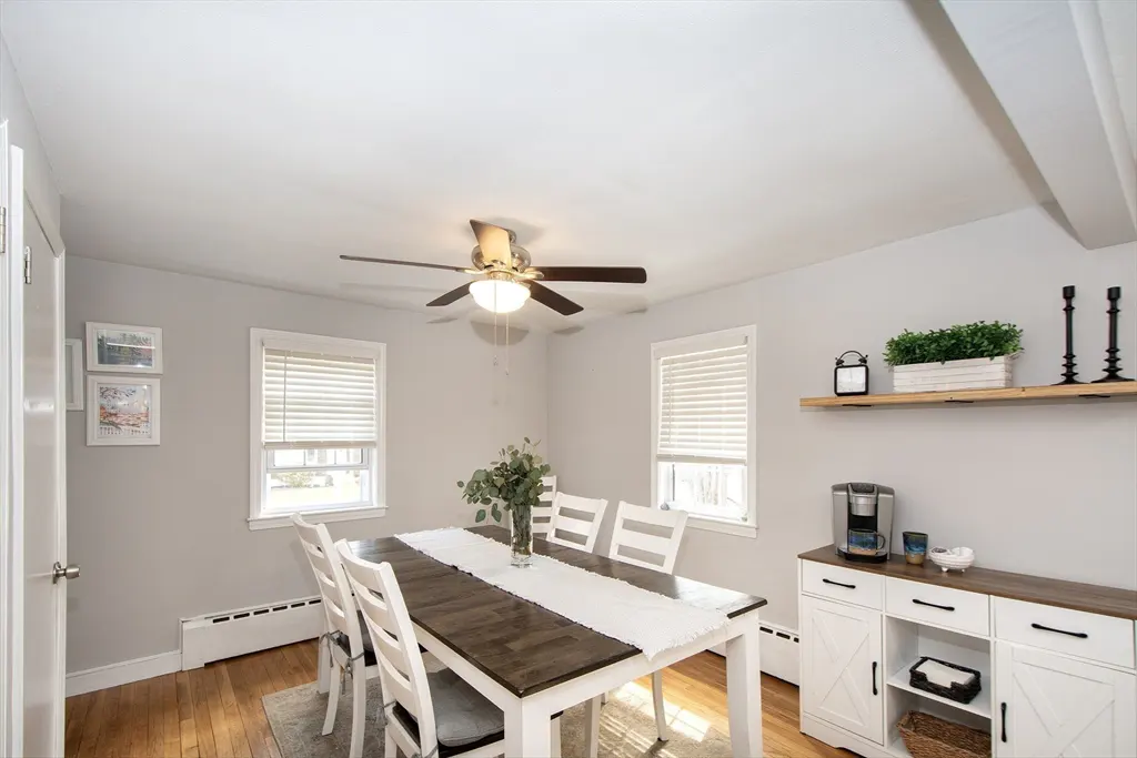 A bright dining room with a dark wood table, white chairs, and a white runner. A ceiling fan hangs above, and a sideboard sits against the wall.