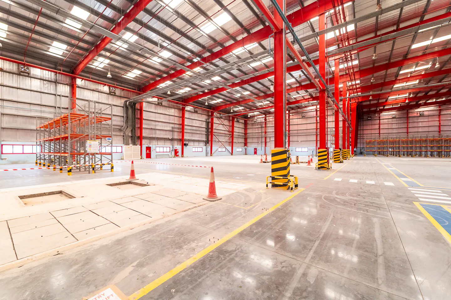 A wide shot of a large, empty warehouse with red support beams and a concrete floor. Shelving units are visible in the background.