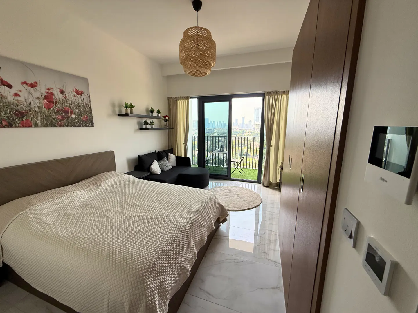 Bedroom with a bed, black sofa, and balcony with city views. A woven pendant light hangs above. A large floral print adorns the wall.