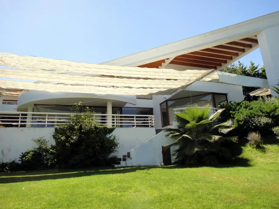Modern white house with a curved balcony and a white mesh sunshade, surrounded by green lawn and trees.
