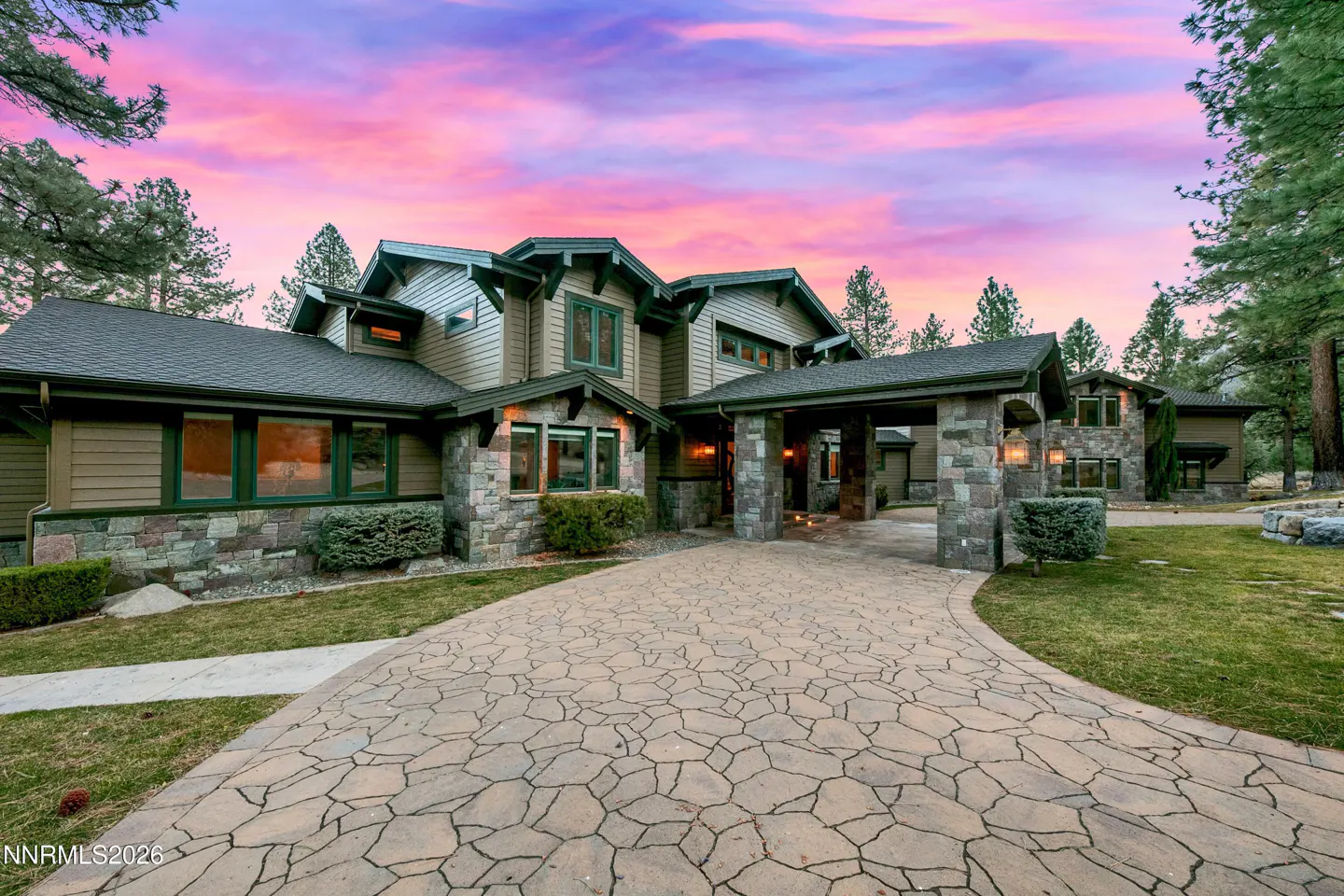 Exterior of a large, two-story home with stone and wood siding, a stone driveway, and a pink and purple sunset sky.