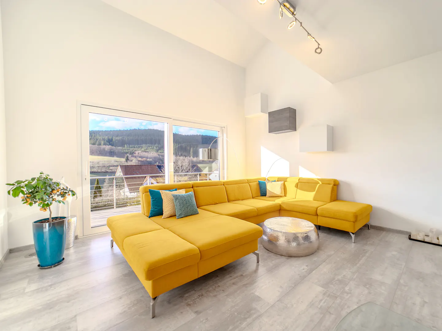 Bright living room with a yellow sectional sofa, blue pillows, and a silver coffee table. Large windows overlook a wooded landscape.