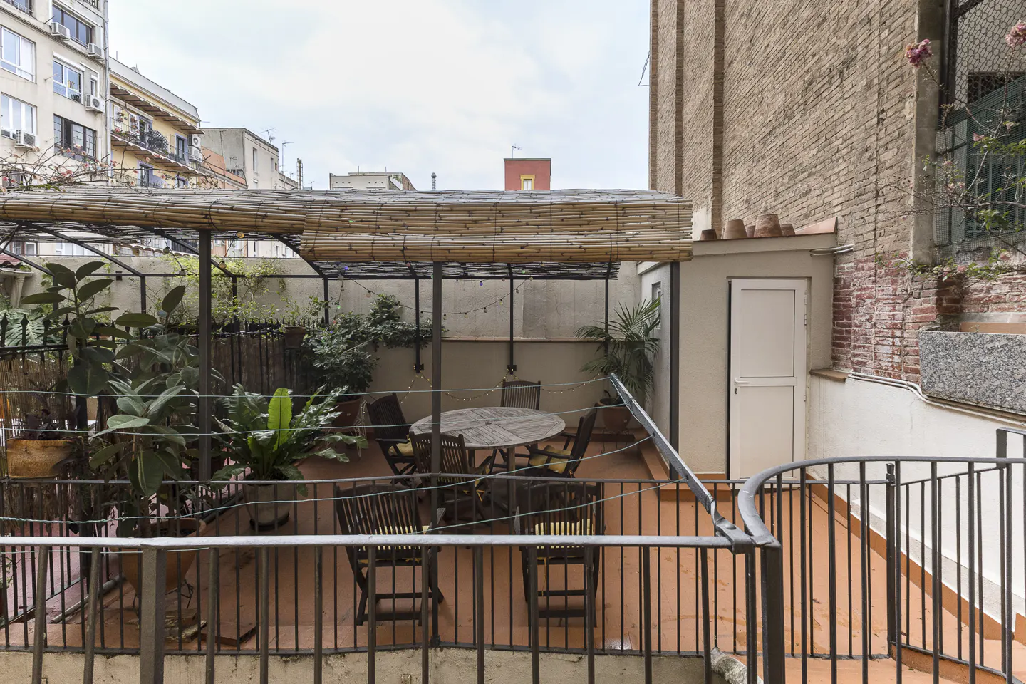 Outdoor terrace with a round wooden table, chairs, potted plants, and a bamboo-covered pergola against a brick building.