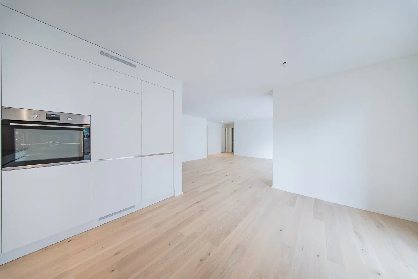 Bright, empty room with light wood floors and white walls. A white kitchen cabinet with a stainless steel oven is on the left.