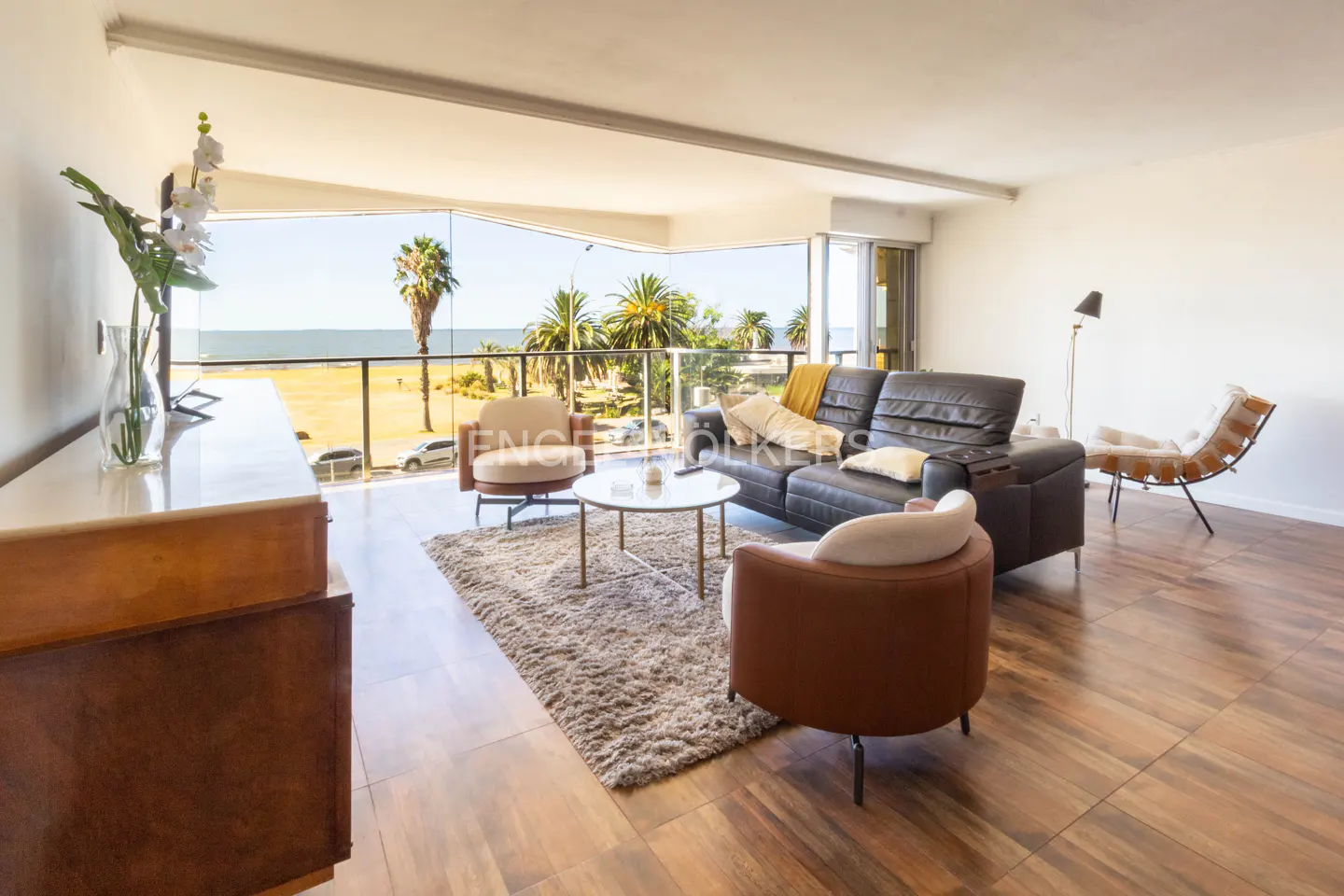 Living room with a brown leather sofa, two armchairs, and a view of the ocean through a glass balcony.