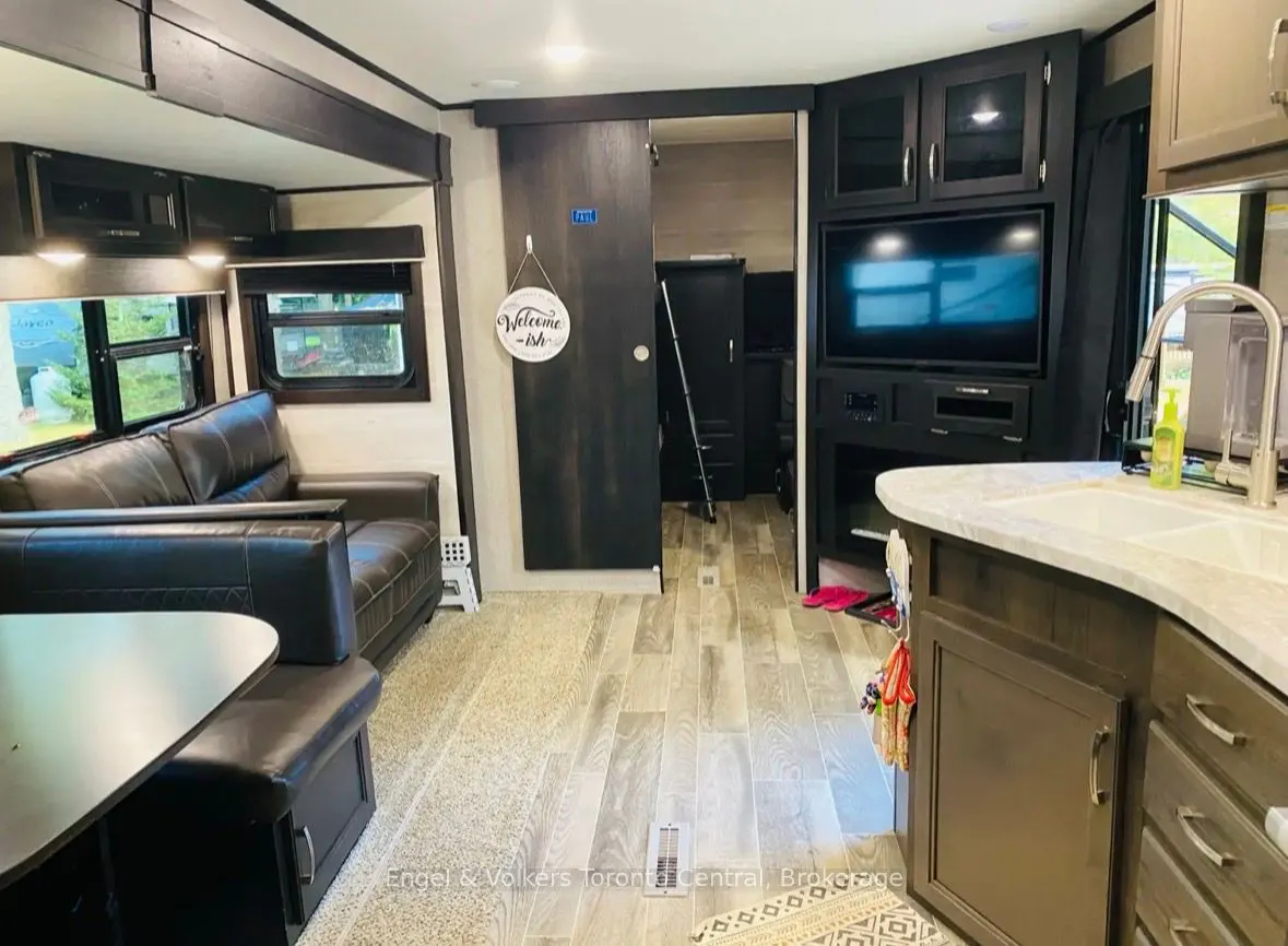 RV interior with a brown leather sofa, table, and a sliding door with a "Welcome-ish" sign. A TV and kitchen area are visible.