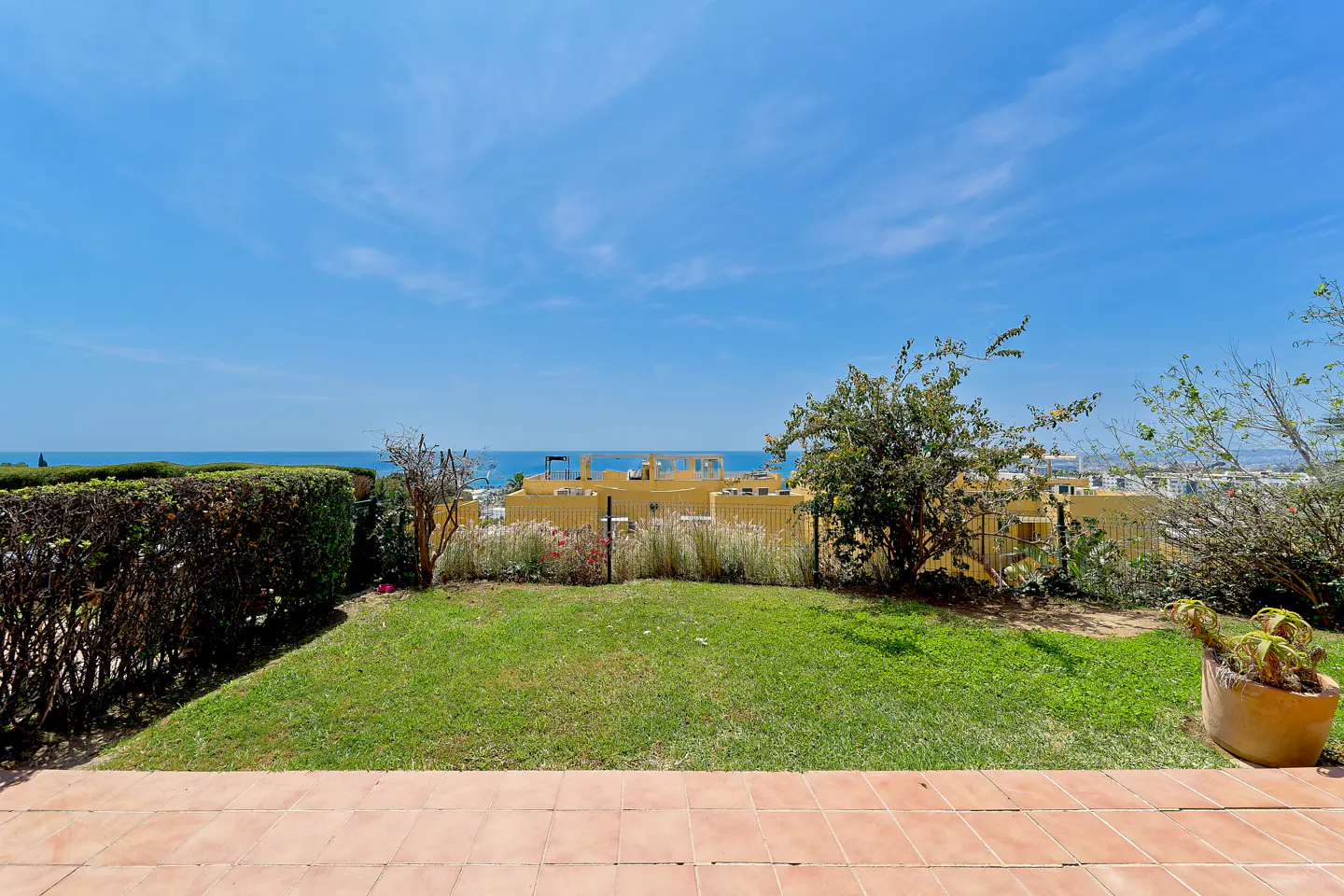 View from a patio with terracotta tiles, a green lawn, and a view of the ocean and blue sky. Bushes and trees surround the lawn.