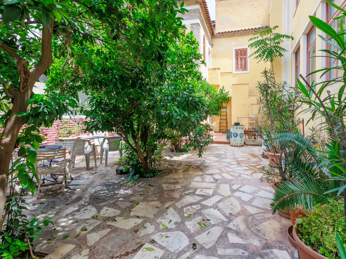 Outdoor courtyard with stone tile flooring, lush green trees, and a yellow building in the background. Patio furniture is visible.