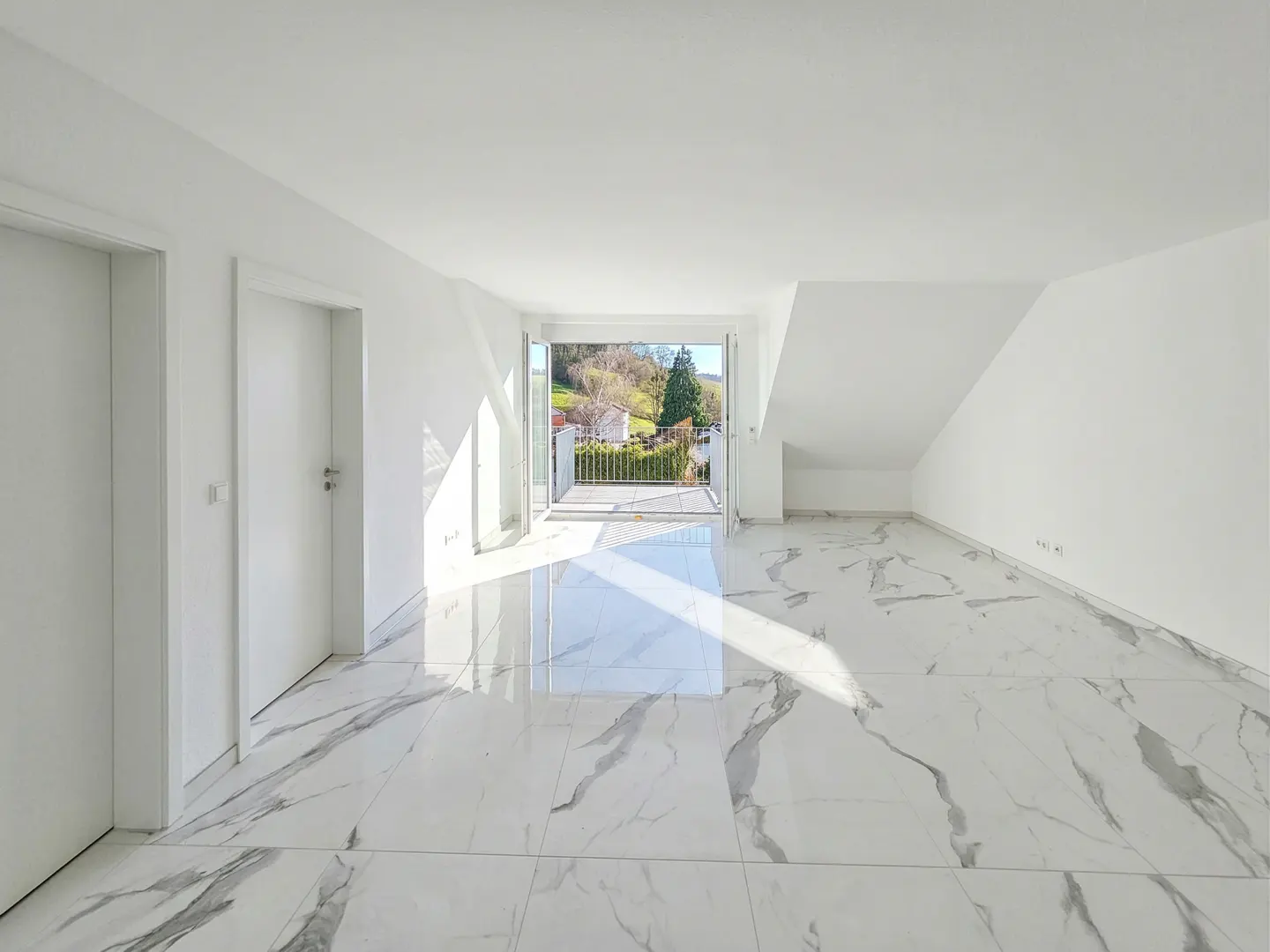 Bright, empty room with white walls and marble floors. Balcony doors open to a view of trees and a house.