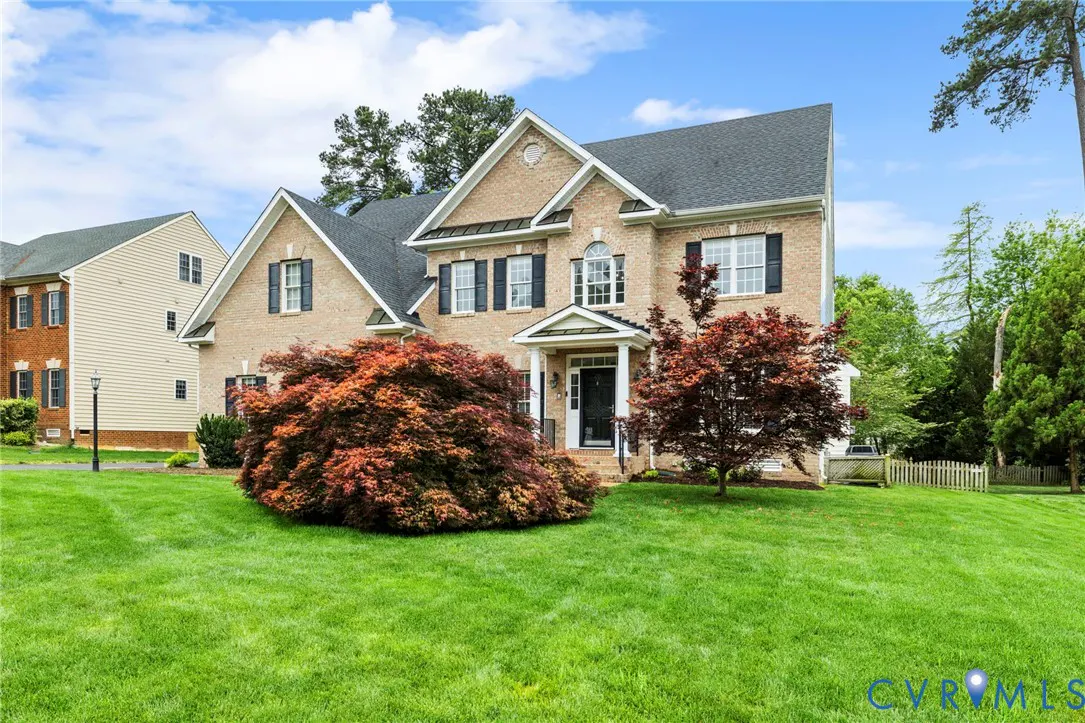Two-story beige brick house with black shutters and a gray roof, surrounded by green lawn and trees.