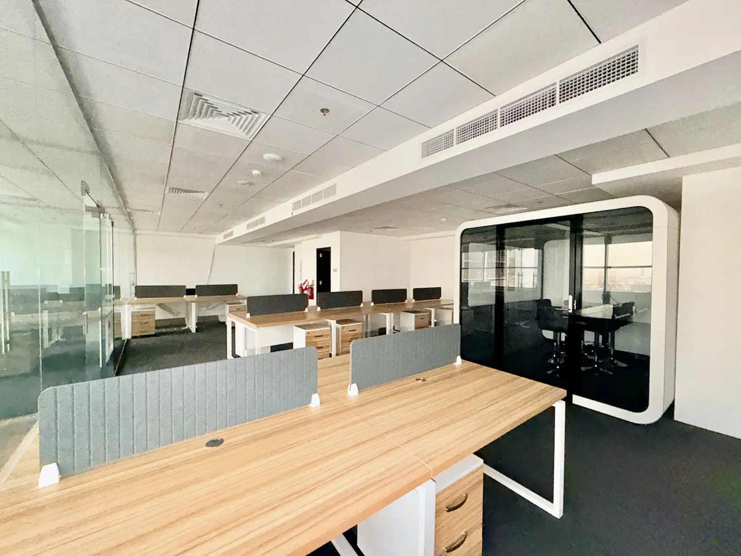 Bright office space with rows of wooden desks, gray dividers, and a modern white soundproof booth with glass walls.