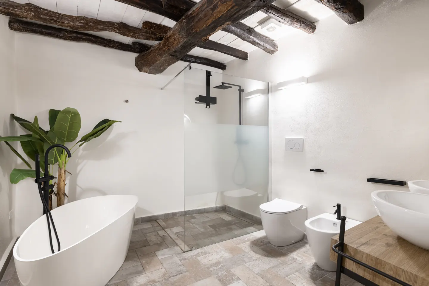 Bright bathroom with white walls, exposed wood beams, and gray tile. Features a tub, glass shower, toilet, bidet, and double sinks.