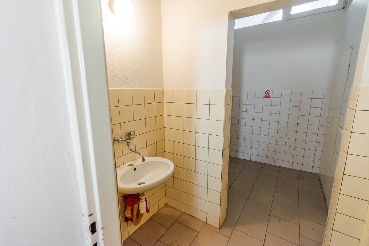 A small washroom with a white wall-mounted sink, cream-colored tiled walls, and a doorway leading to a tiled restroom area.