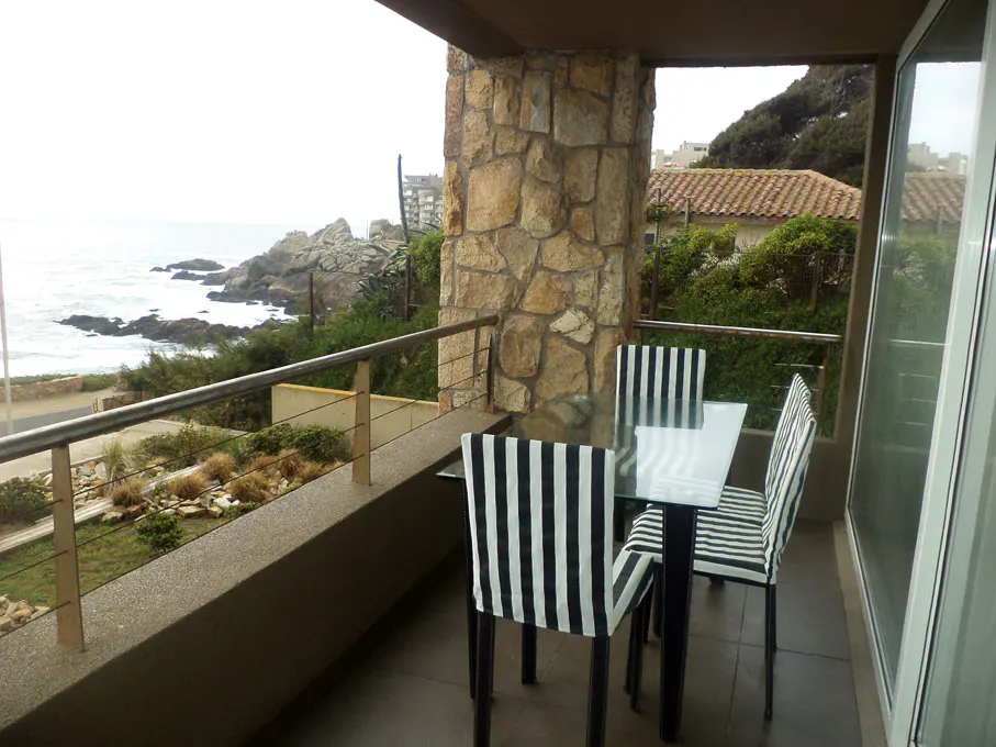 Balcony with ocean view features a glass table and striped chairs. Stone pillar and metal railing add architectural detail.