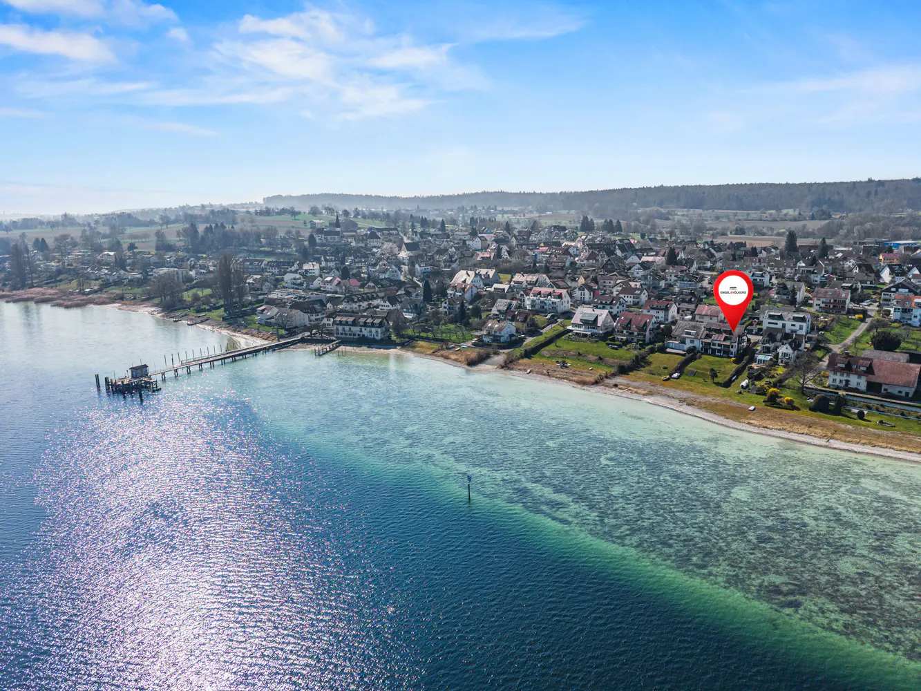 Aerial view of a lakeside town with a red pin marking a property. Blue water, green hills, and houses visible.