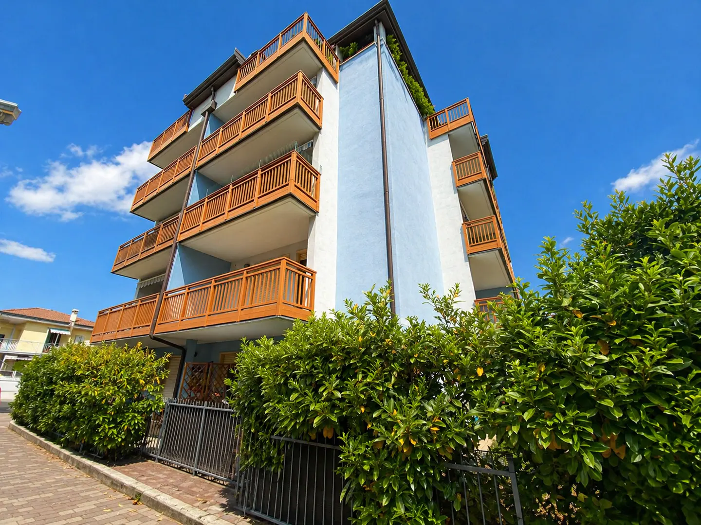 Low angle view of a light blue apartment building with wood balconies under a blue sky. Green bushes line the front.