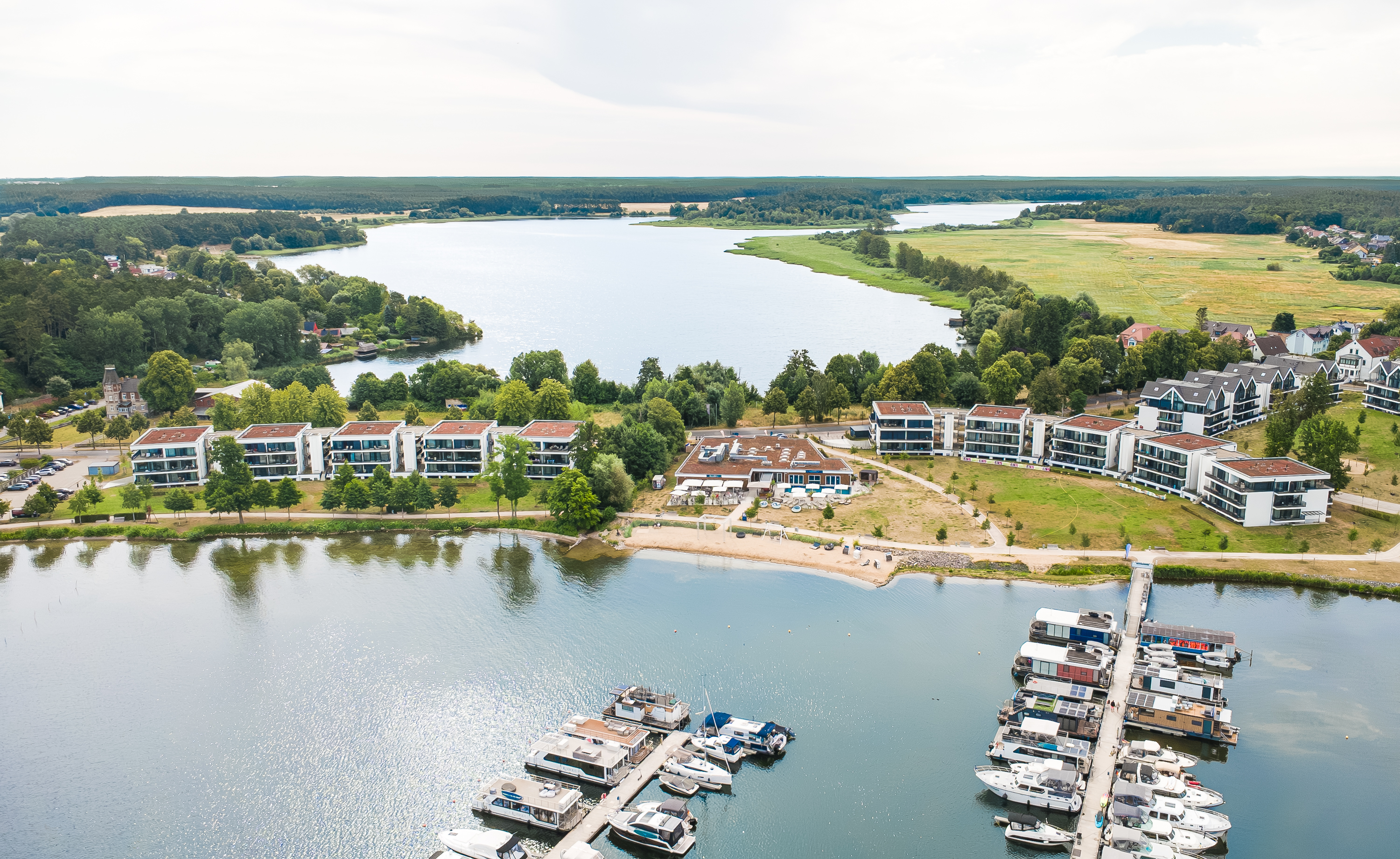 Aerial view of modern apartment buildings along a lake with a marina full of boats and a small sandy beach.