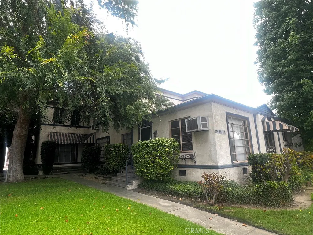 Two-story stucco house with green lawn, trees, and striped awnings. An air conditioner is visible in a window.