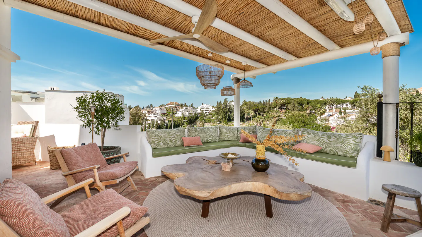 Outdoor patio with a thatched roof, wicker lights, and a large wooden table. Seating includes chairs and a built-in bench with patterned cushions.