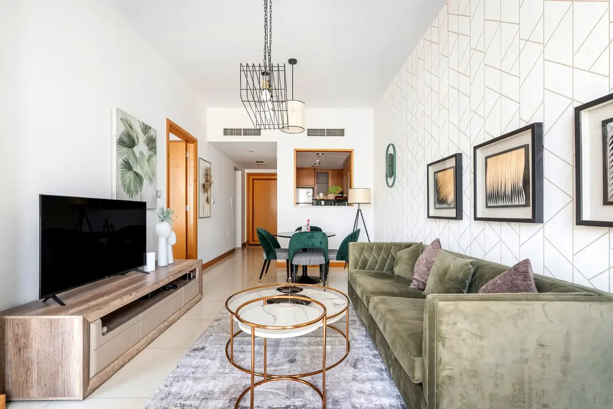 A modern living room with a green velvet sofa, round marble tables, and a dining area with green chairs. Geometric wallpaper adds a stylish touch.