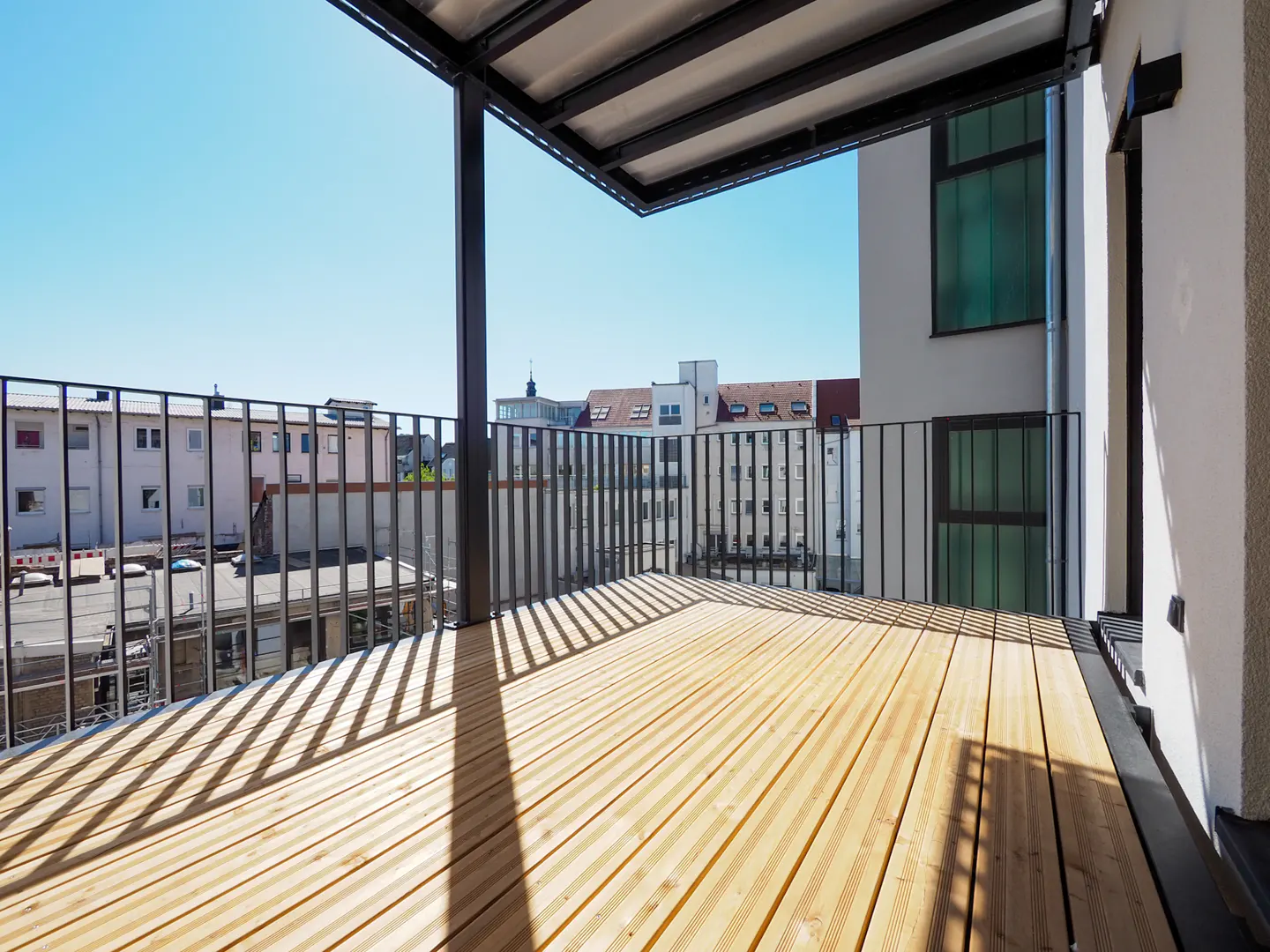 Modern balcony with light wood decking, black metal railings, and a partial roof, overlooking a sunny urban cityscape.