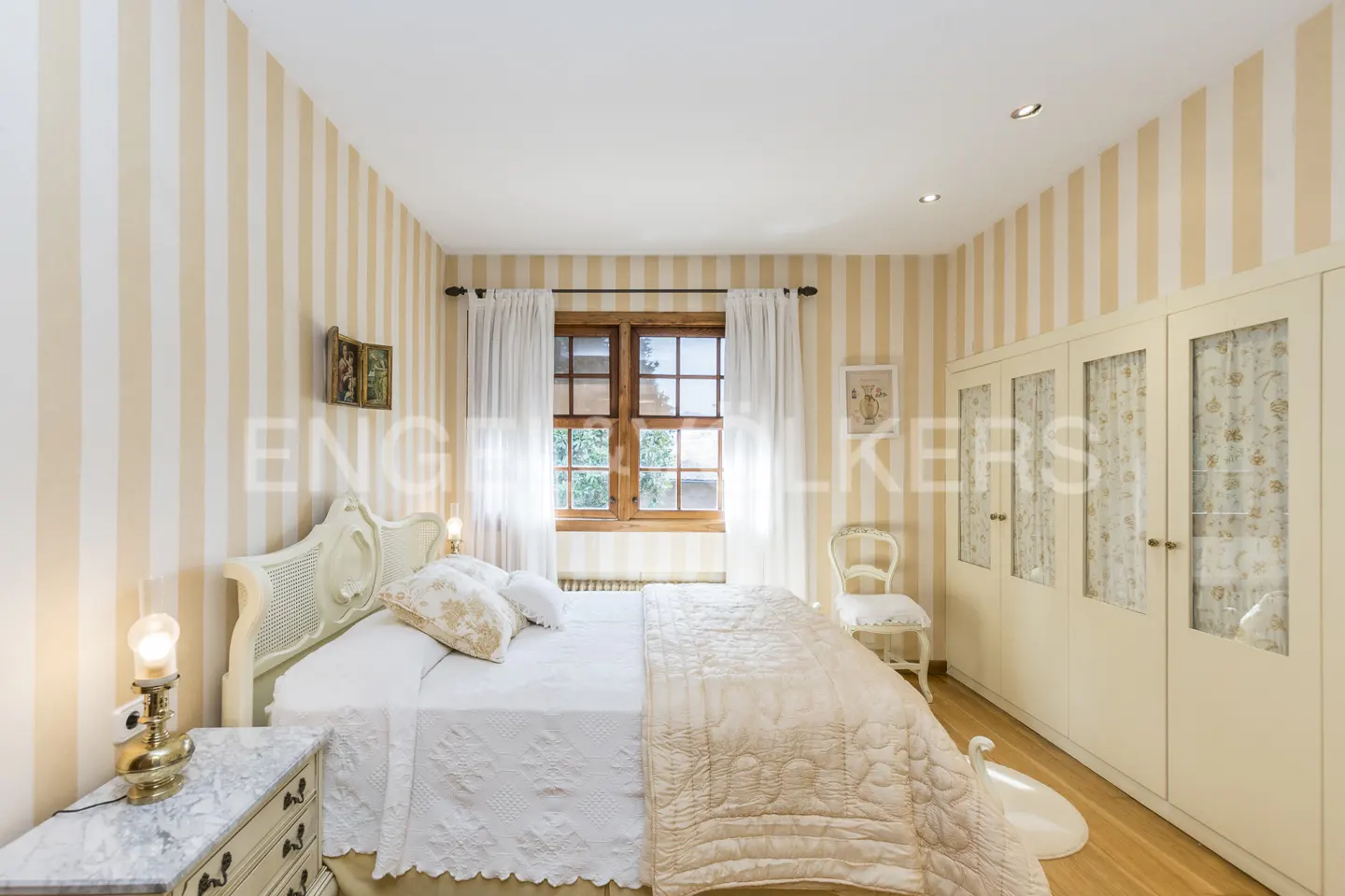 Bedroom with striped wallpaper, white bed, wooden window frame, and cream-colored wardrobe.