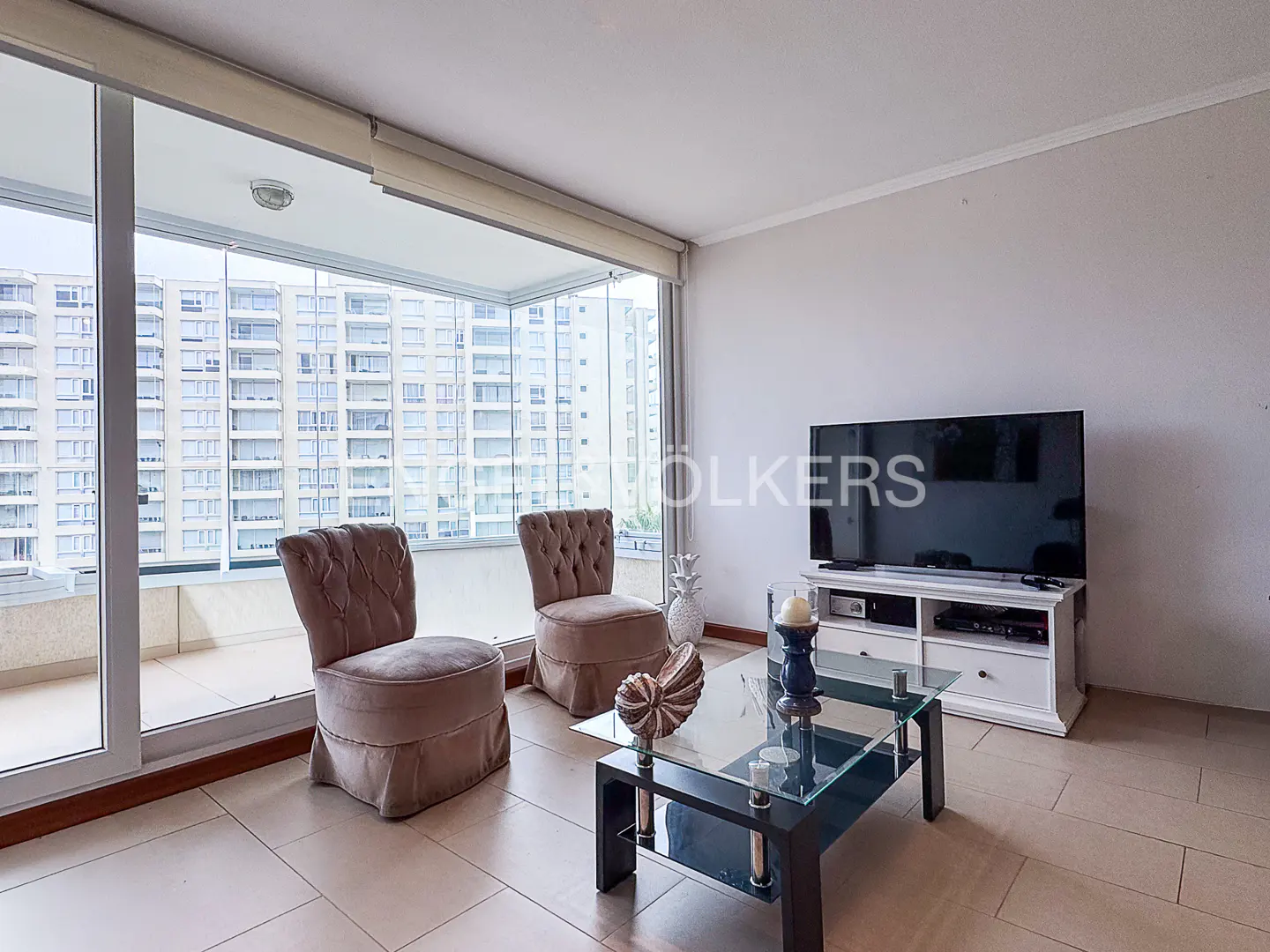 Living room with two tufted chairs, glass coffee table, TV, and balcony with city view.