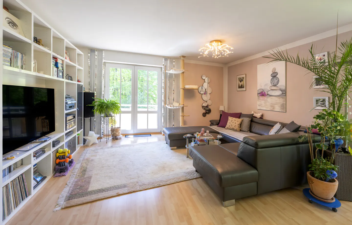 Living room with a brown leather sectional sofa, white shelves, and a large TV. A light rug covers the wood floor. Plants and art decorate the space.