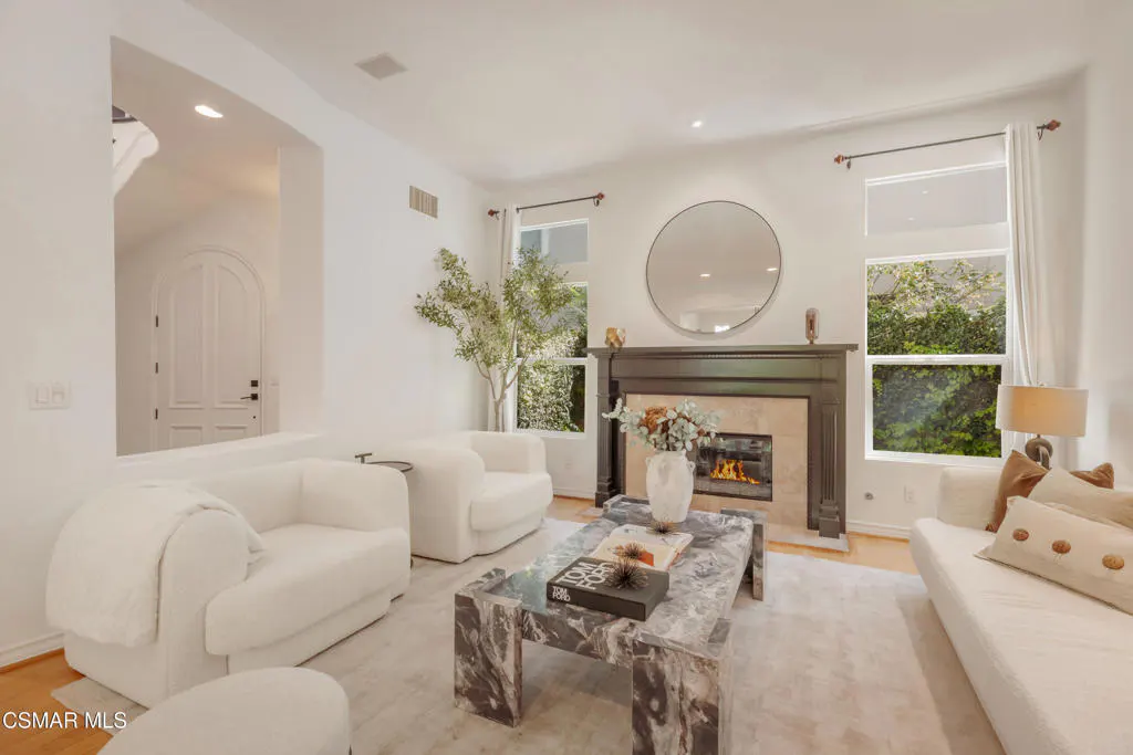 Bright living room with white walls, chairs, and sofa. A marble coffee table sits on a light rug. A fireplace with a round mirror above it adds a focal point.