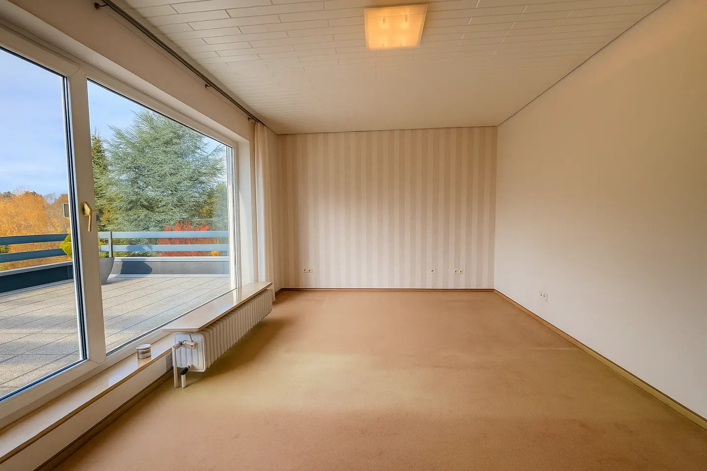 Bright, empty room with beige carpet, striped wallpaper, and a large window to a balcony with trees.
