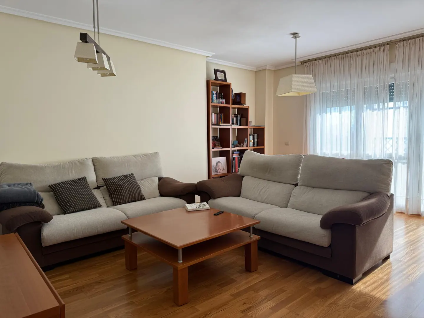Living room with two sofas, a wooden coffee table, and a bookshelf. Sunlight streams through sheer curtains.