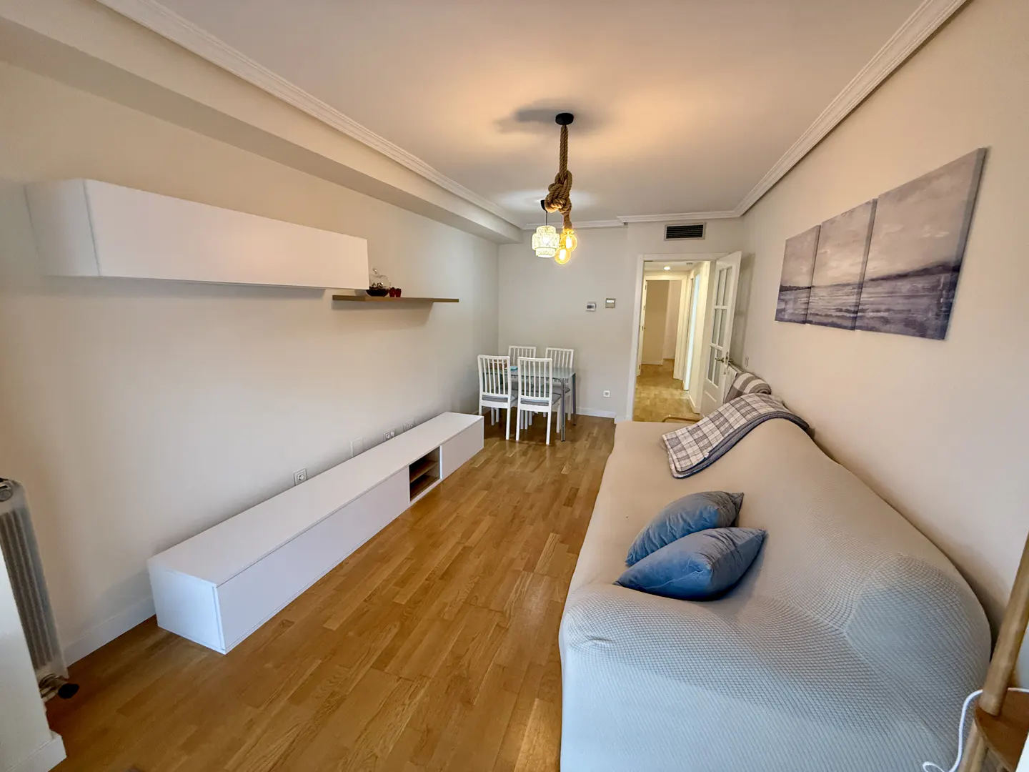 Living room with hardwood floors, a beige sofa with blue pillows, and a white dining set. A nautical-themed light fixture hangs from the ceiling.