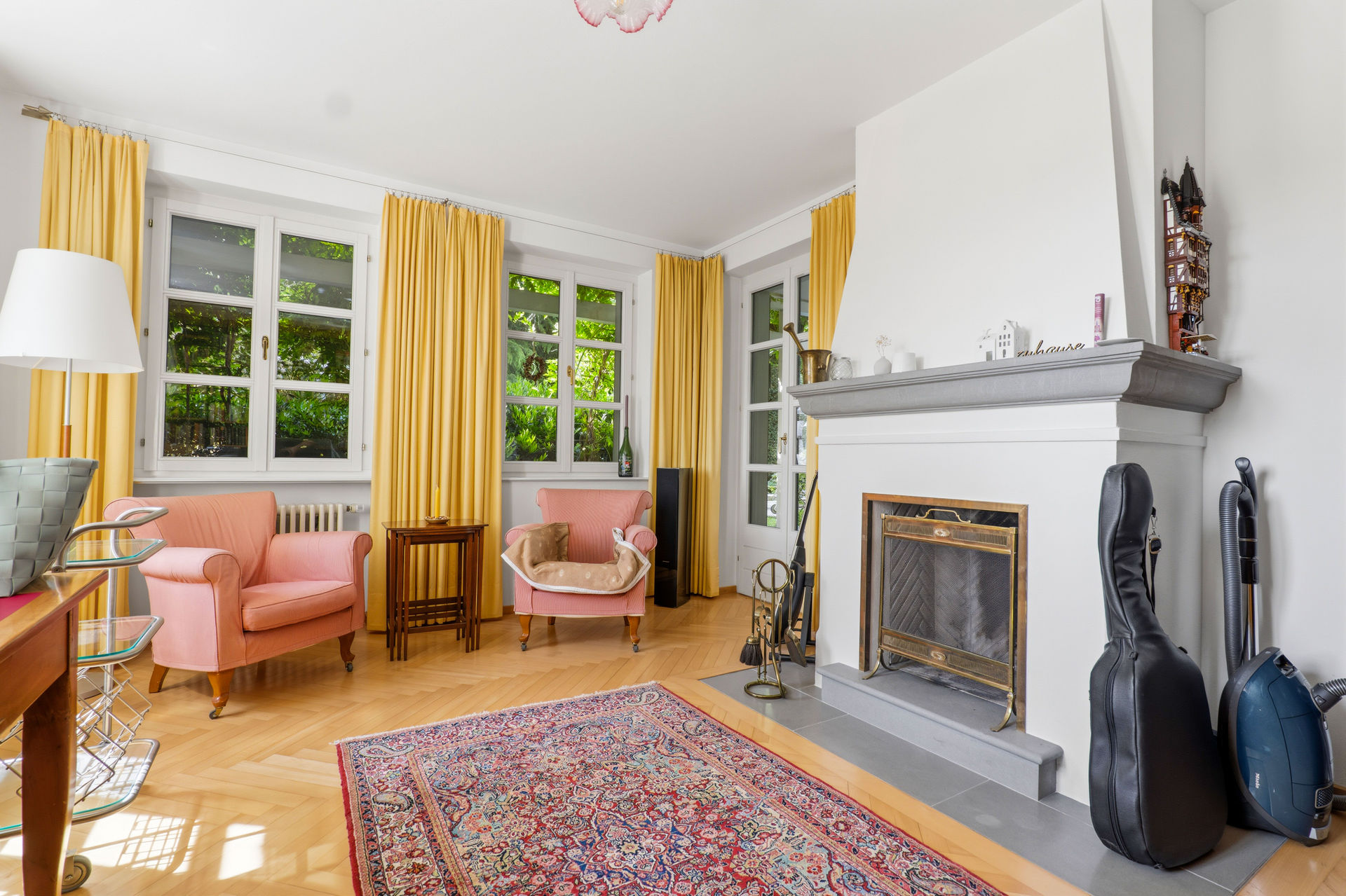 Living room with two pink armchairs, yellow curtains, a patterned rug, and a white fireplace with a black guitar case. [AI-generated text]