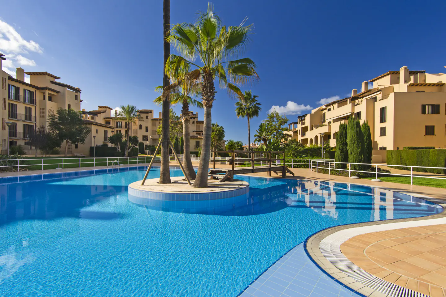 A bright blue swimming pool with a palm tree island, surrounded by tan buildings under a clear blue sky.