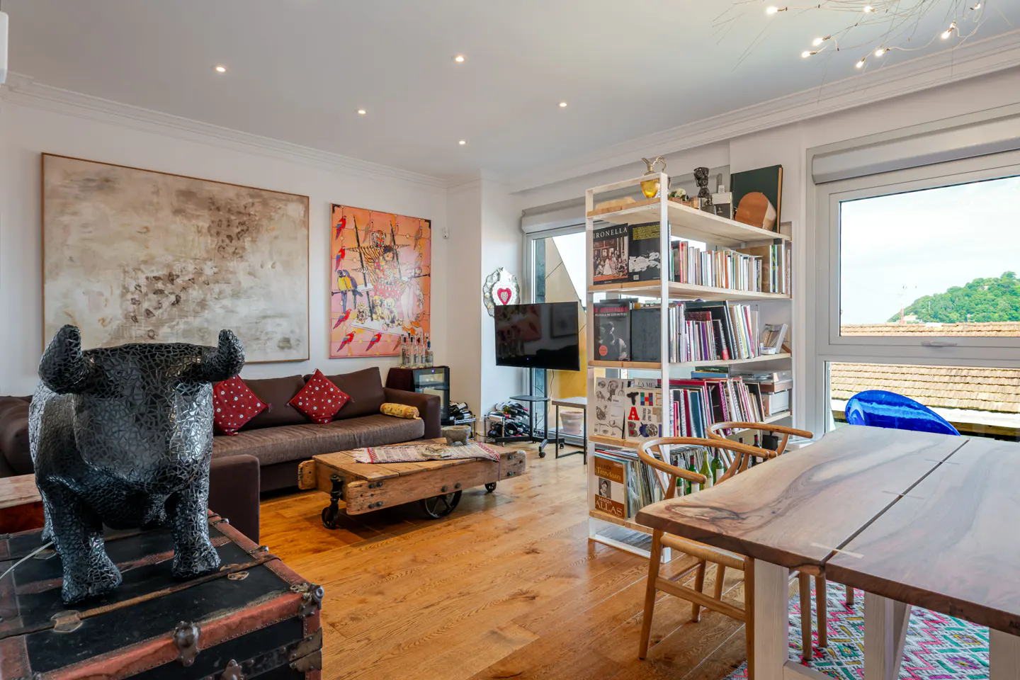 Living room with wood floors, brown sofa, and a black bull statue on a trunk. Bookshelf, table, and large paintings decorate the space.
