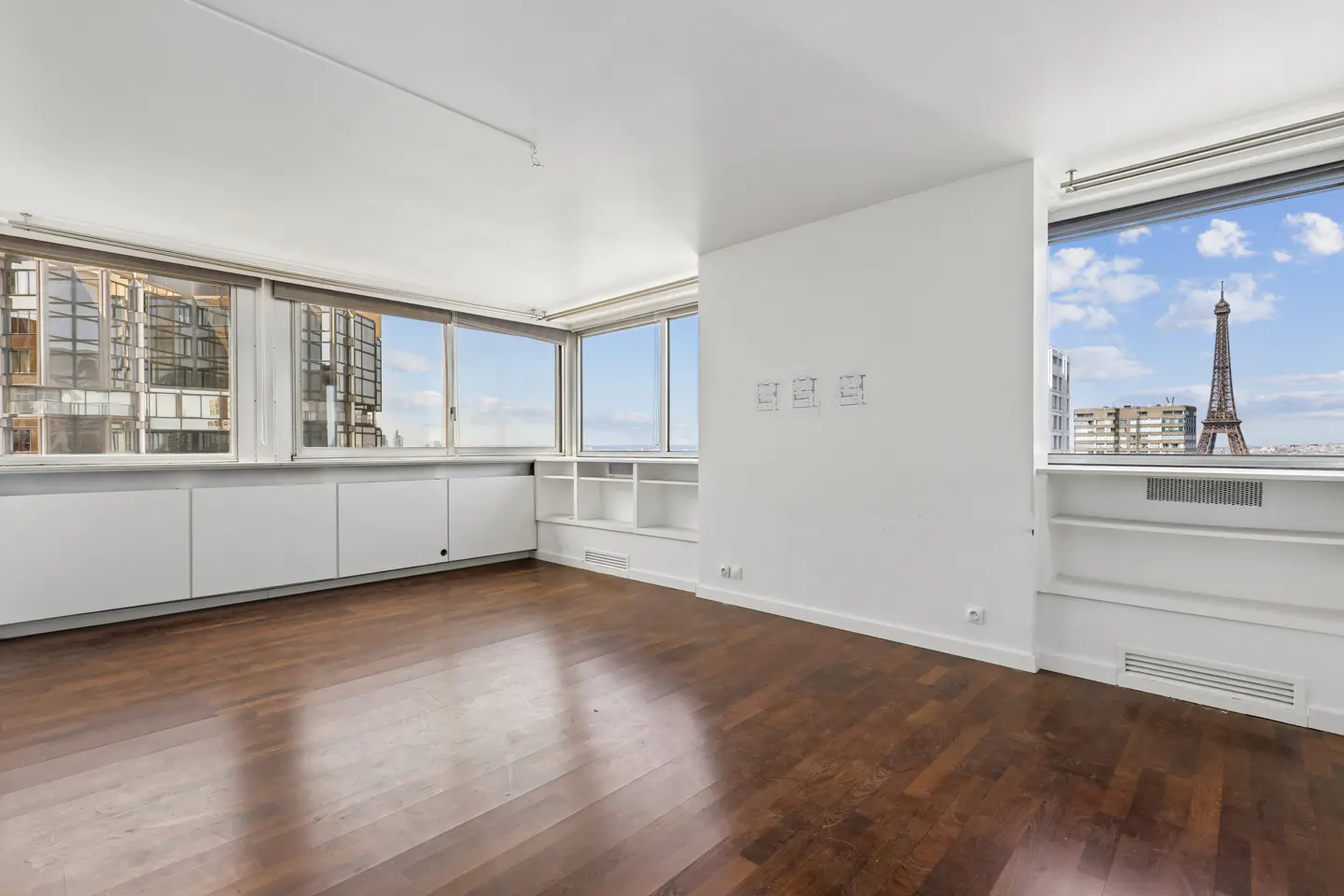 Bright, empty room with hardwood floors and white walls. Large windows offer a view of the Eiffel Tower.