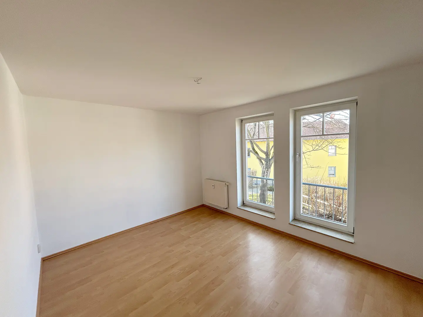Empty room with white walls, light wood flooring, a radiator, and two windows overlooking a yellow building.