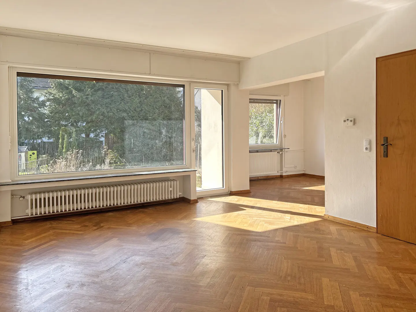 Bright, empty room with herringbone wood floors, white walls, and a large window overlooking green trees. A wood door is on the right.