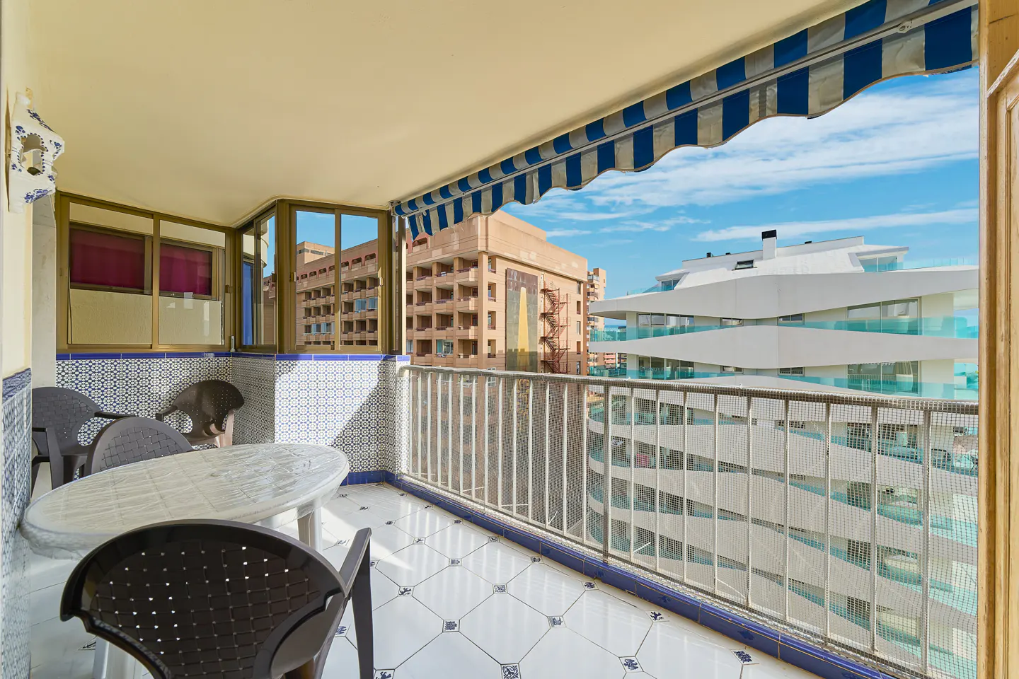 Covered balcony with a white table and chairs, blue and white tiled walls, and a view of nearby apartment buildings.