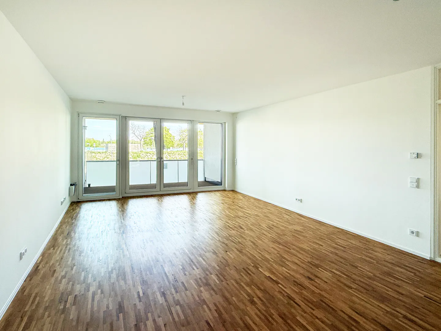 Bright, empty room with hardwood floors and white walls. Glass doors lead to a balcony with a view of trees.