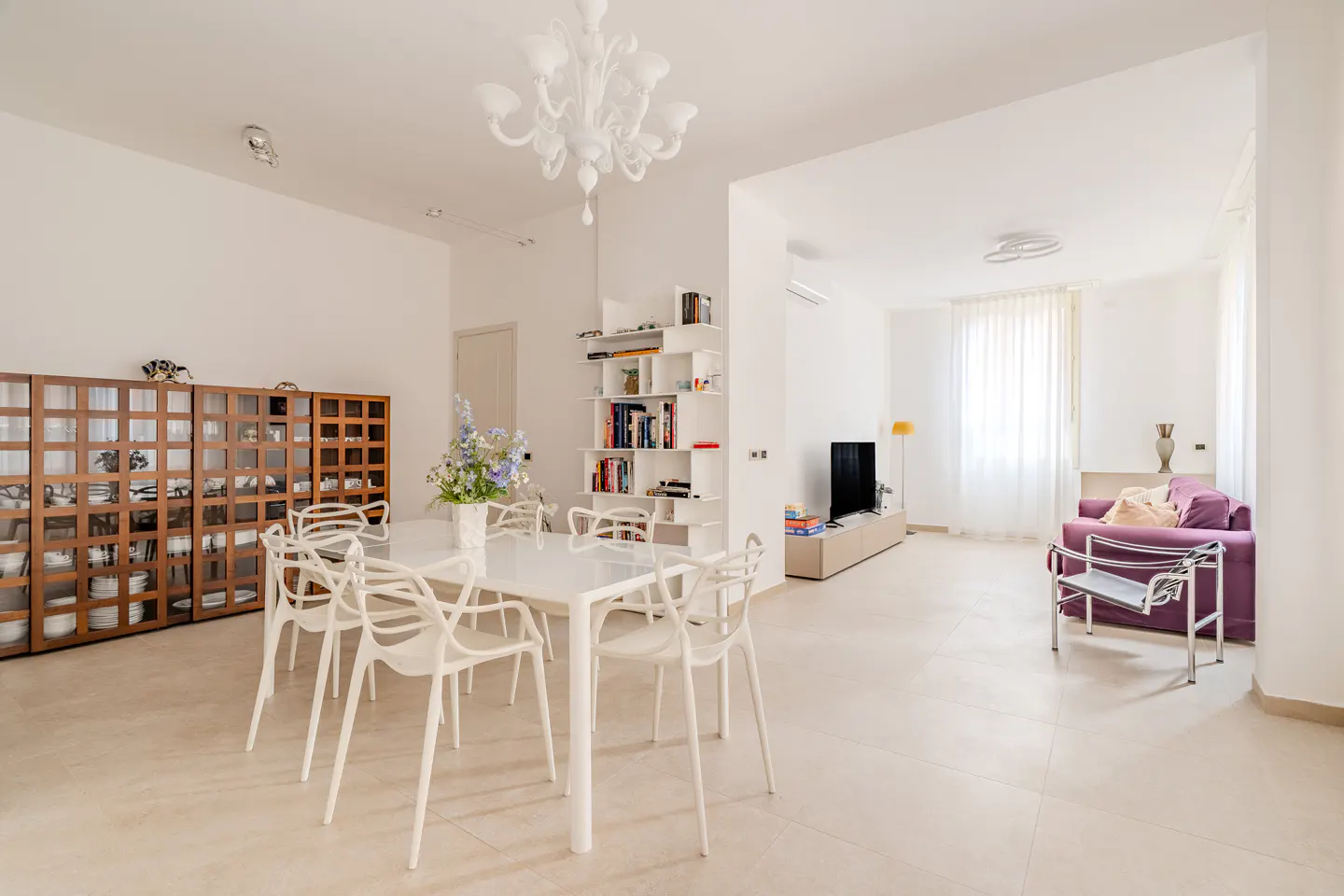 Bright, open-concept living and dining area with white walls, tile floors, and modern furnishings. A white dining table with chairs sits under a chandelier.