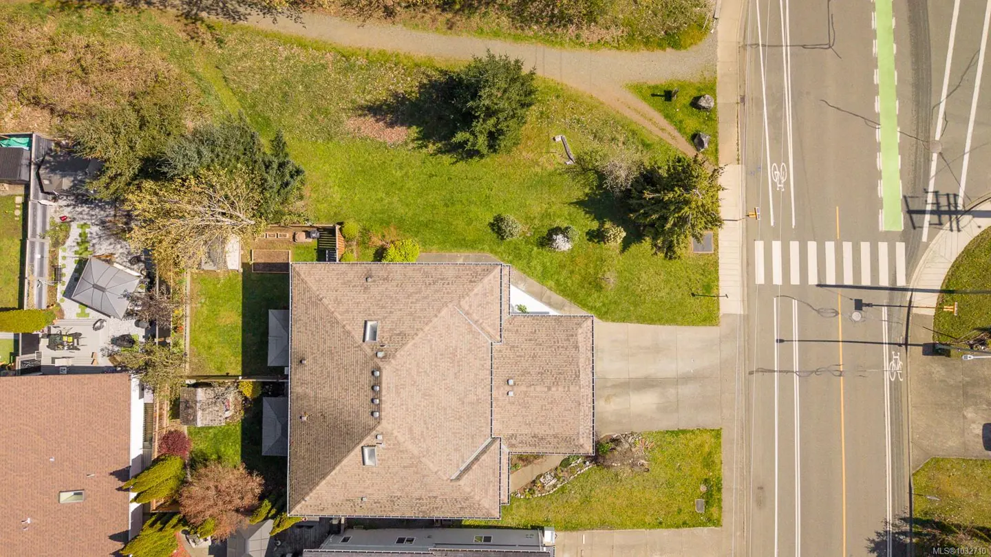 Aerial view of a brown-roofed house with a driveway, lawn, and trees next to a road with a crosswalk and bike lane.
