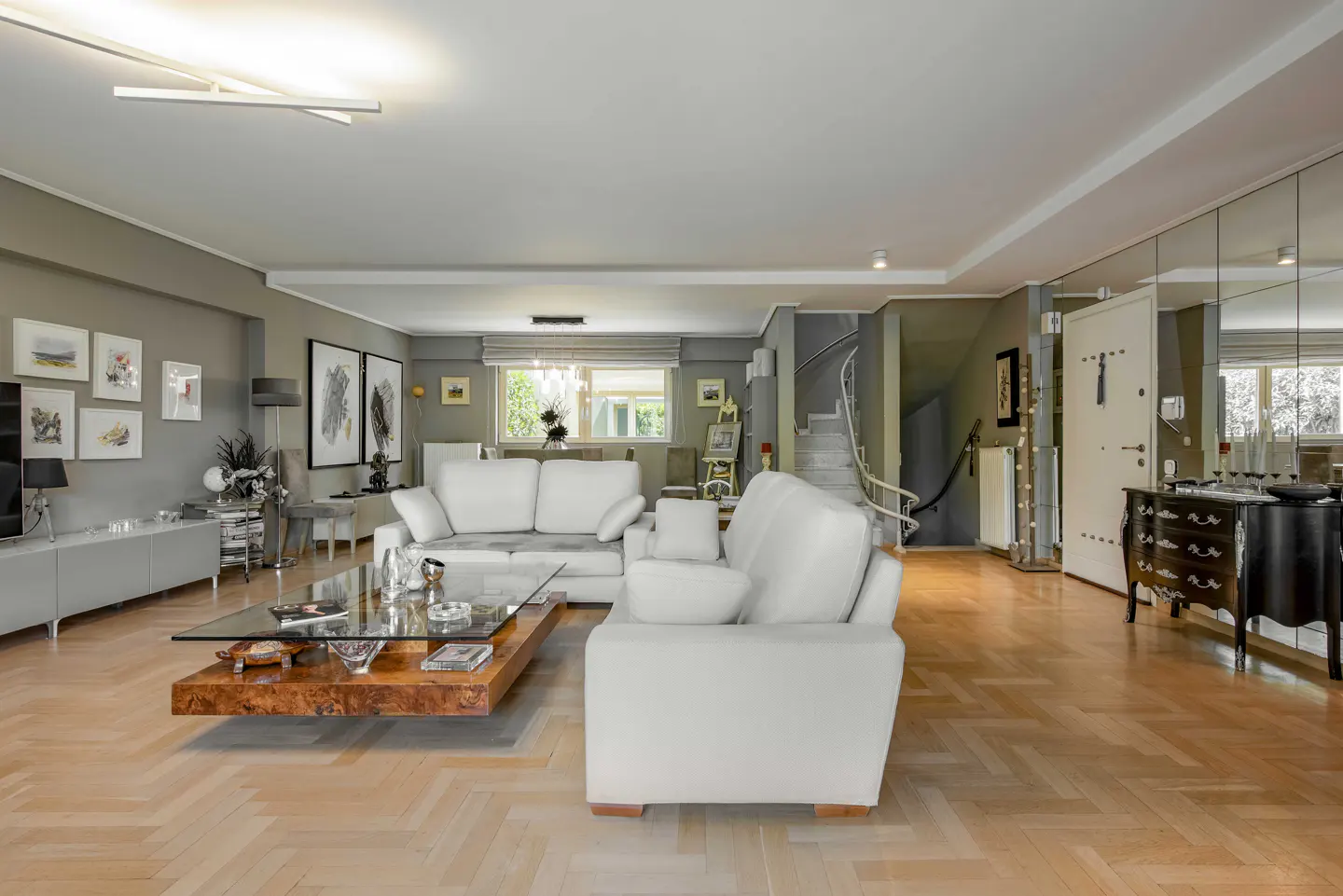 A modern living room with white sofas, a wood and glass coffee table, and herringbone wood floors. Art adorns the gray walls.